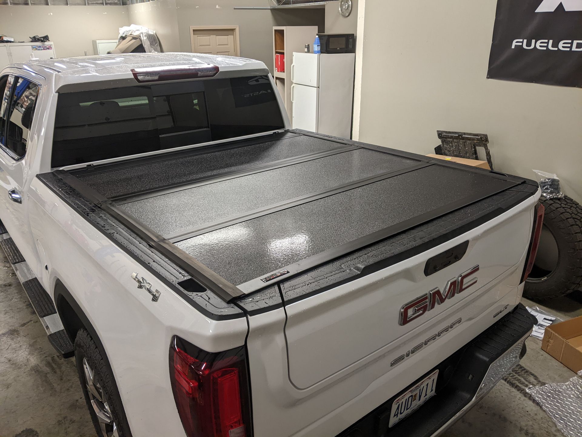 White GMC pickup truck with a black tonneau cover parked indoors in a garage or showroom.