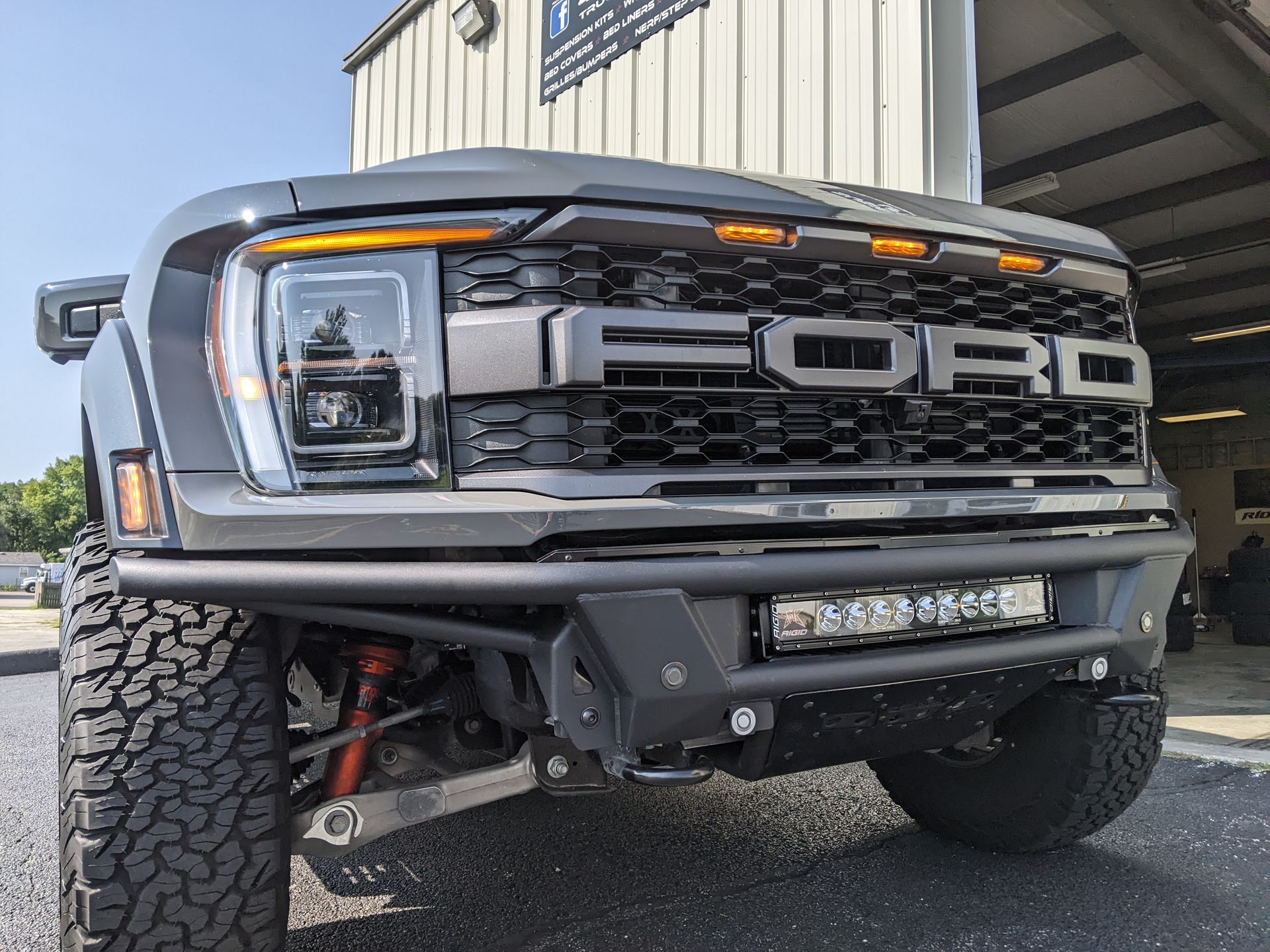 Gray lifted Ford Raptor front end parked by a metal building, showing grille and off-road tires