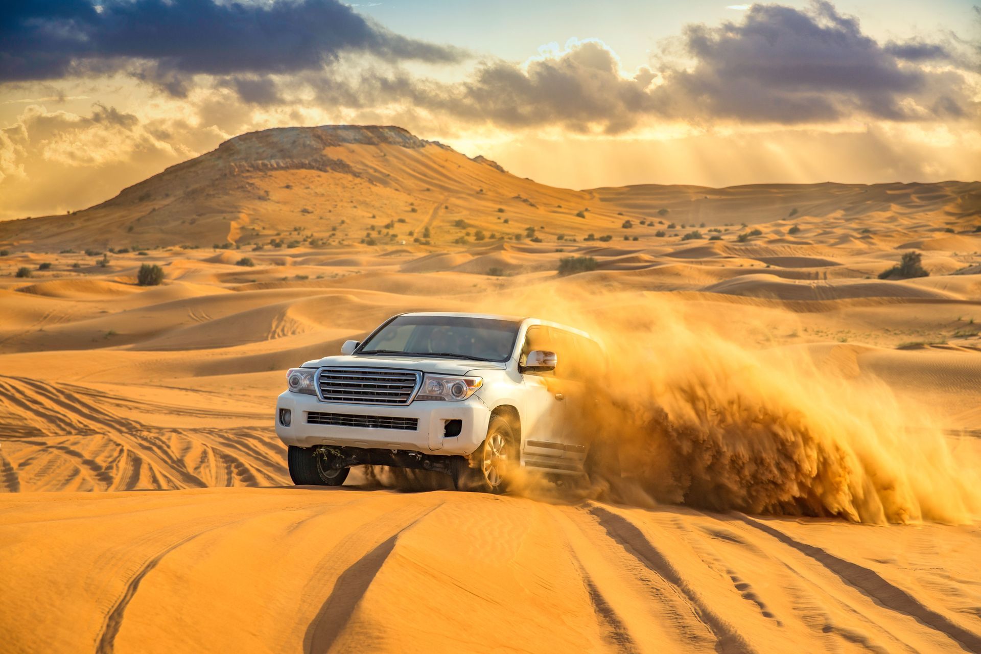 White SUV drifting through desert sand dunes at sunset, kicking up a cloud of sand