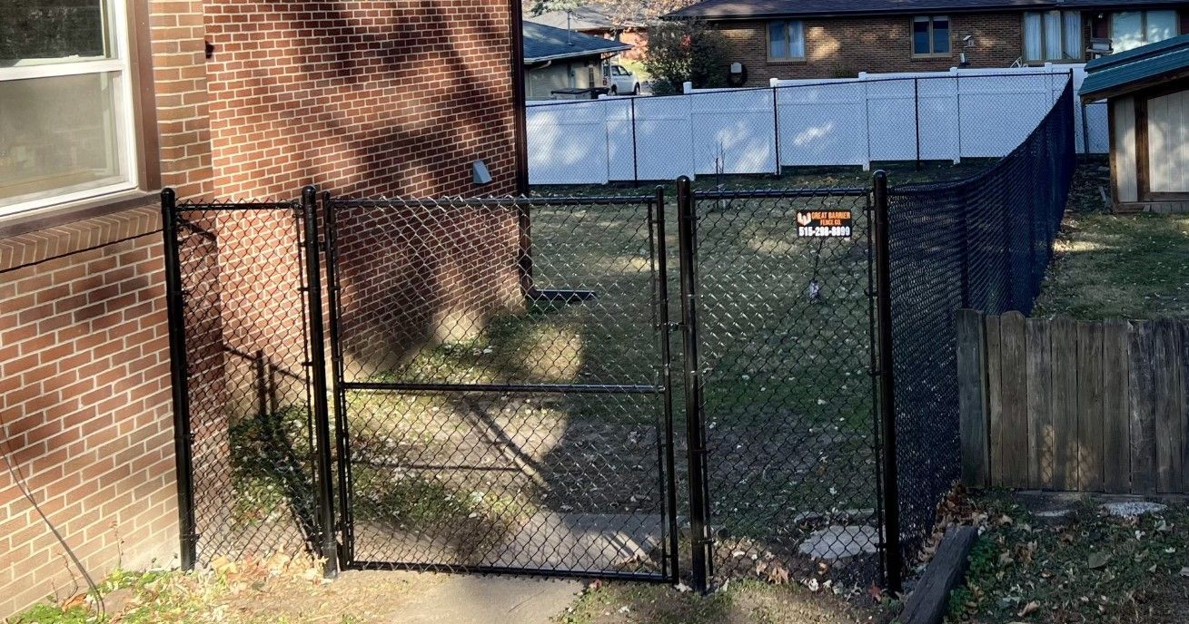 A black chain-link gate attached to a brick building, opening into a fenced backyard with grass and scattered leaves.