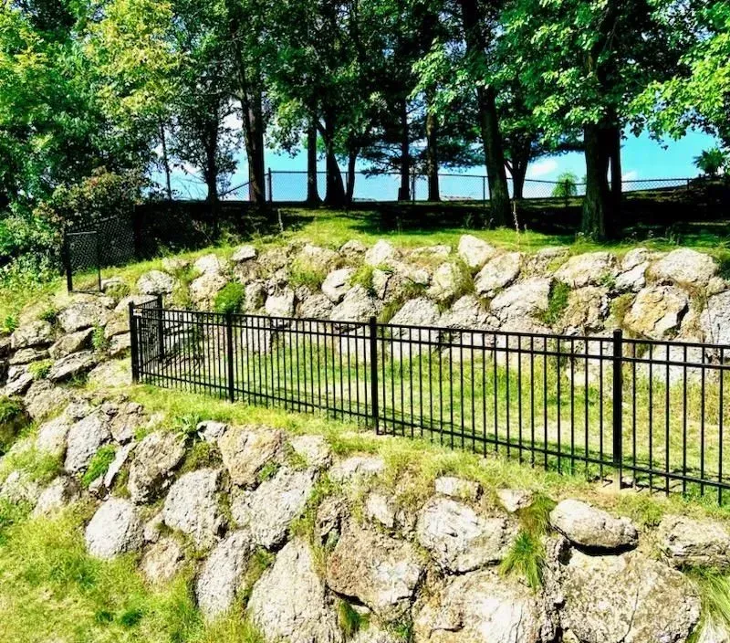 Black fence on a rocky hillside with trees and a lake in the background. Green grass and blue sky.