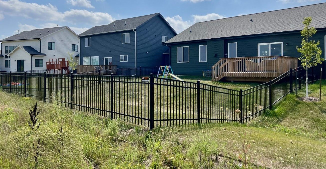 A black metal fence encloses backyard spaces behind several modern suburban houses on a sunny day.