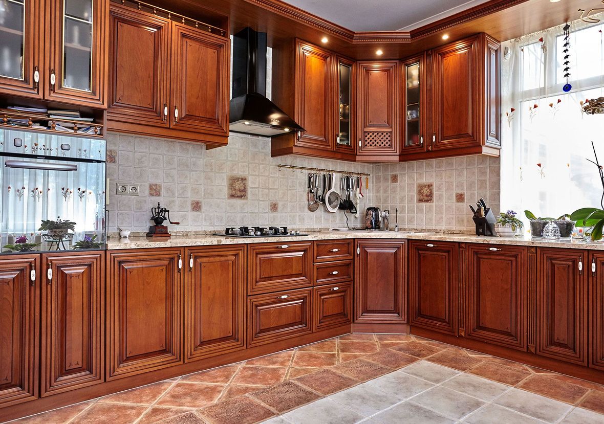 Warm wood kitchen with L-shaped cabinets, tiled backsplash, black range hood, and a sunlit window.