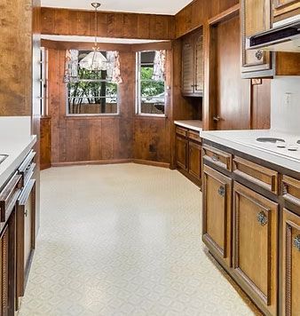 Wood-paneled kitchen with white countertops and a light patterned floor, viewed toward a windowed dining nook