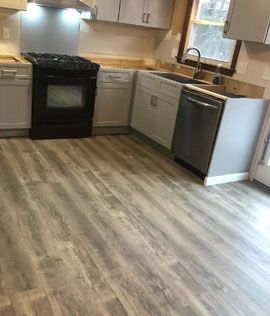 Empty kitchen with gray wood flooring, black stove, white cabinets, and a dishwasher by a window.