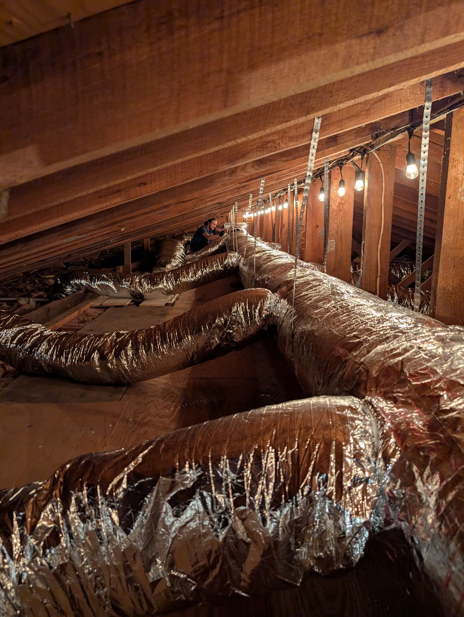 Air ducts snaking across an attic with wood beams and string lights.