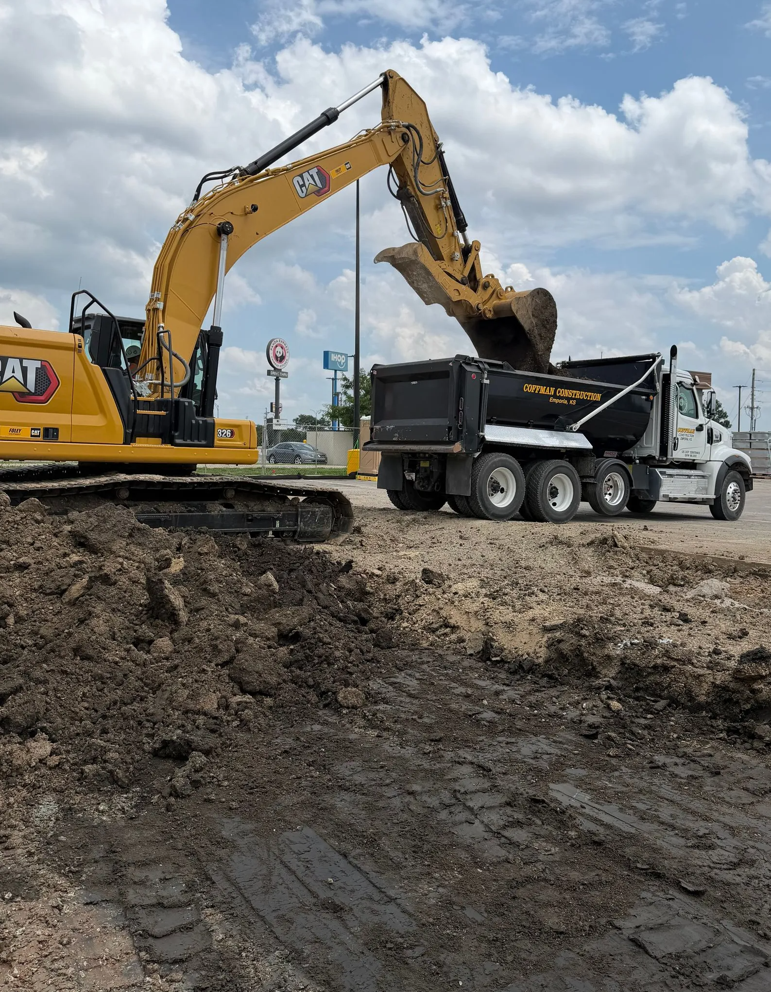 Yellow excavator loading dirt into a black dump truck on a muddy construction site under a cloudy sky