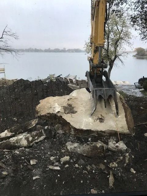 Excavator digging into a rocky shoreline beside a calm body of water under an overcast sky