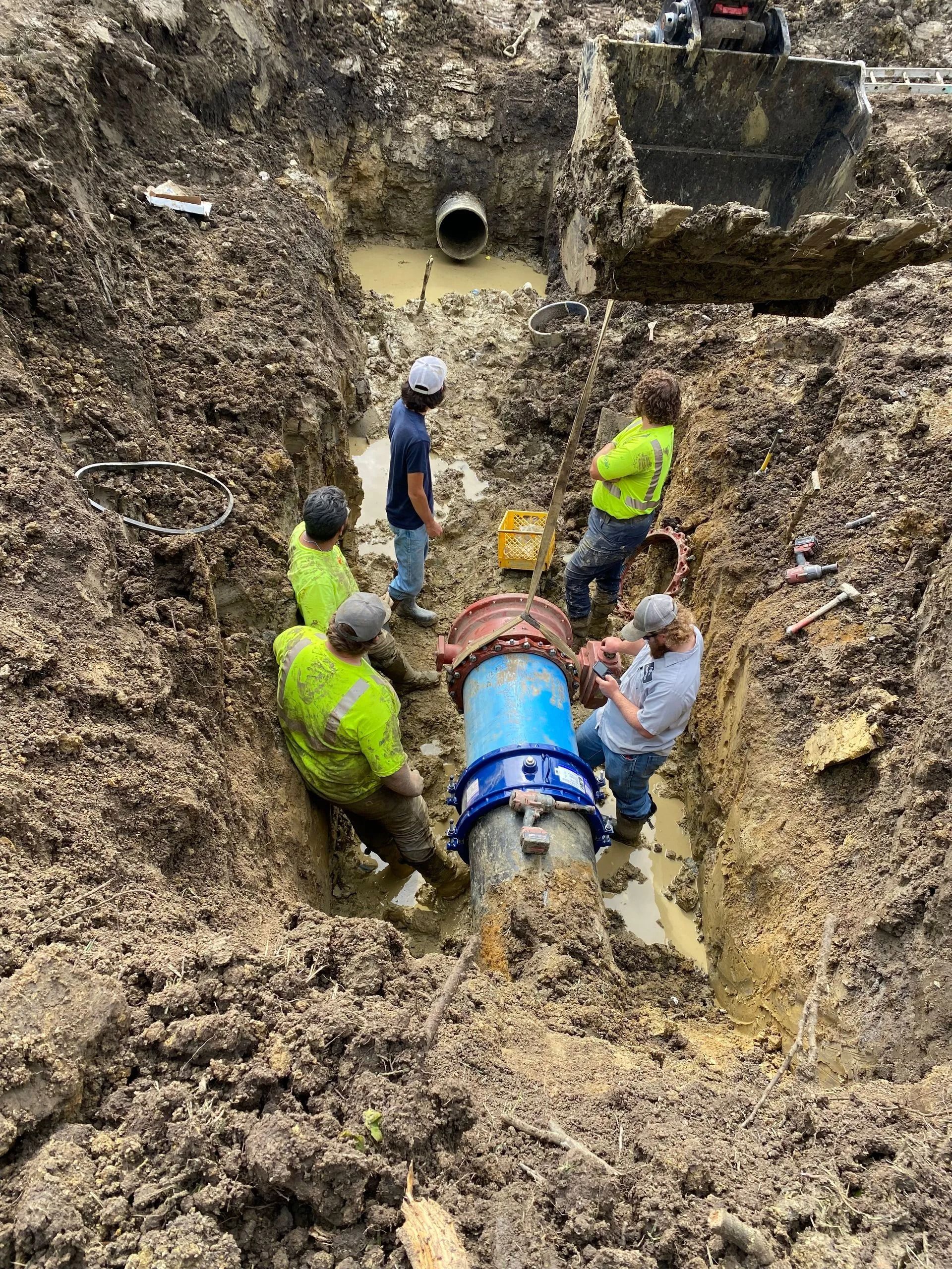 Workers repair a blue pipe in a muddy trench, surrounded by dirt walls and excavation equipment.