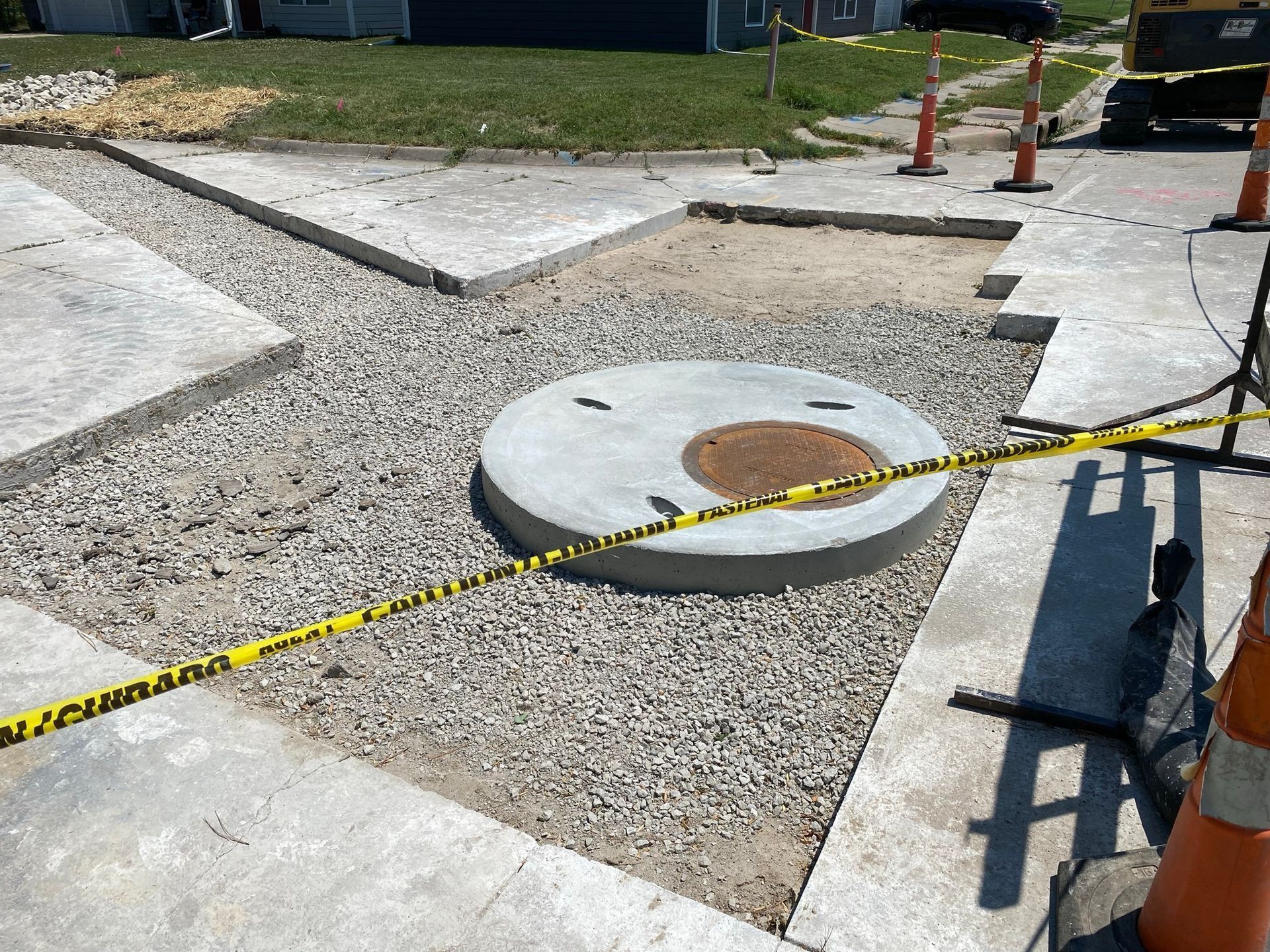 Construction site with a round concrete manhole cover surrounded by gravel and caution tape.