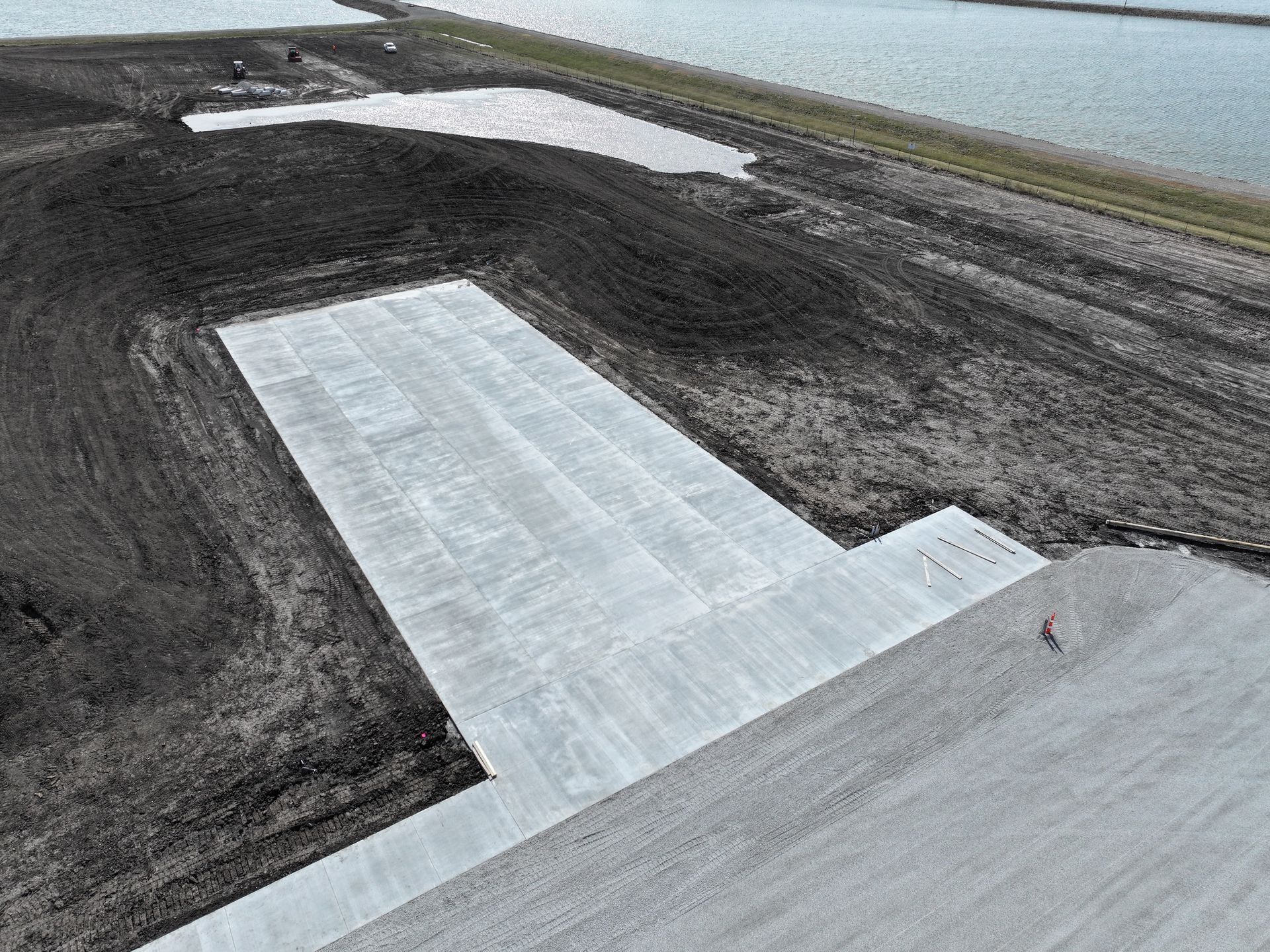 Aerial view of a large paved concrete pad beside dark gravel and a body of water.