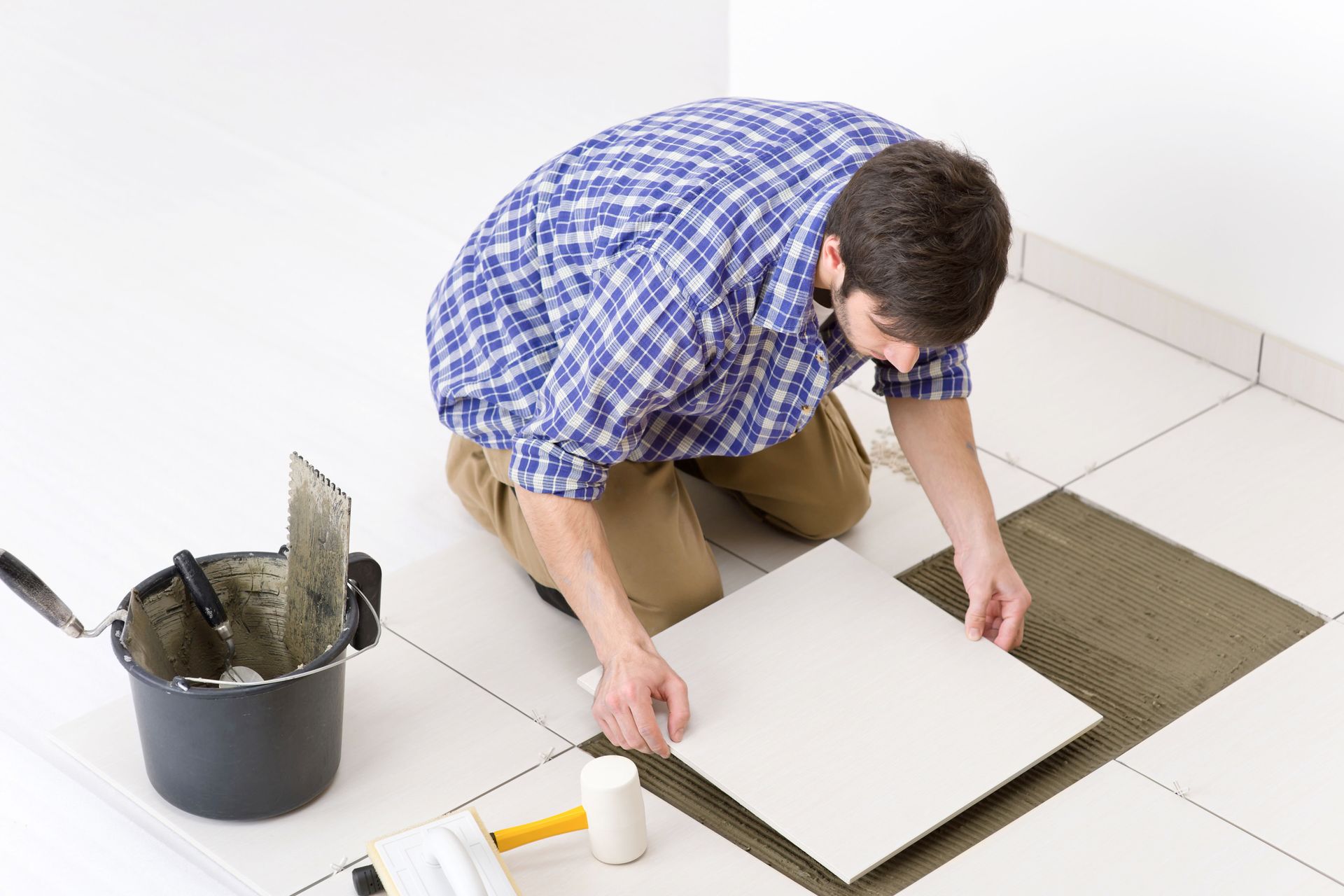 Man kneeling on white tile, installing a floor tile with tools and bucket nearby