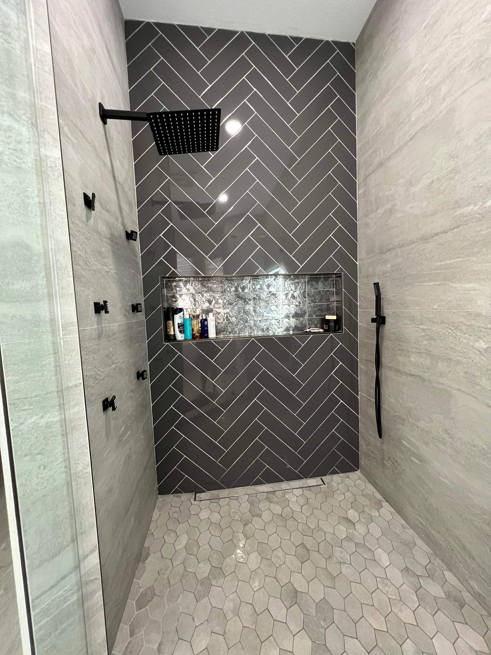 Gray tiled shower with herringbone accent wall, pebble floor, multiple shower heads.