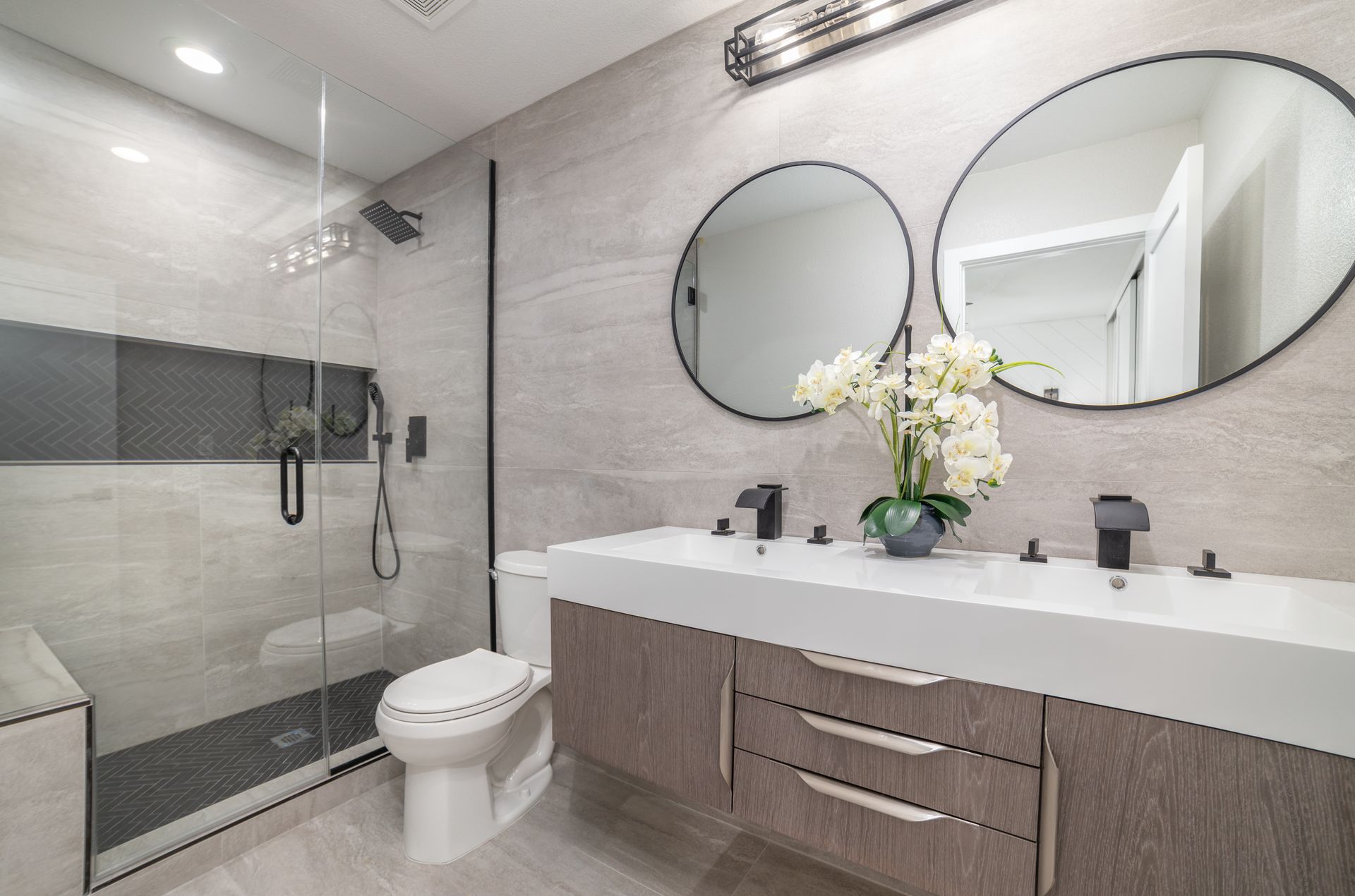 Modern bathroom with double sink, round mirrors, glass shower, and gray tile walls.