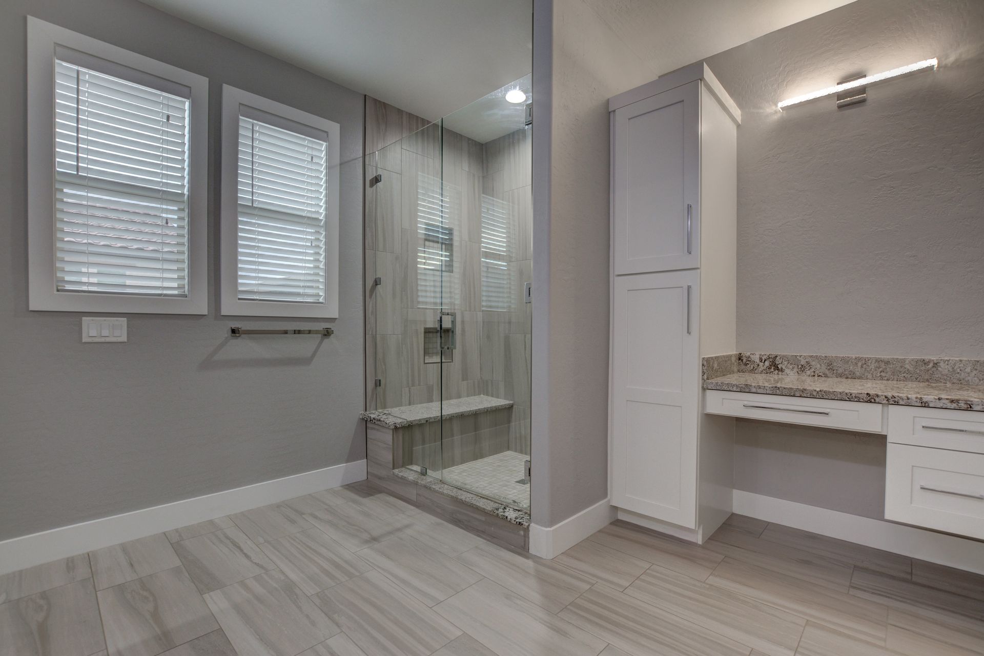 Bathroom with gray walls, wood-look floor, glass shower, white cabinets, and two windows with blinds.