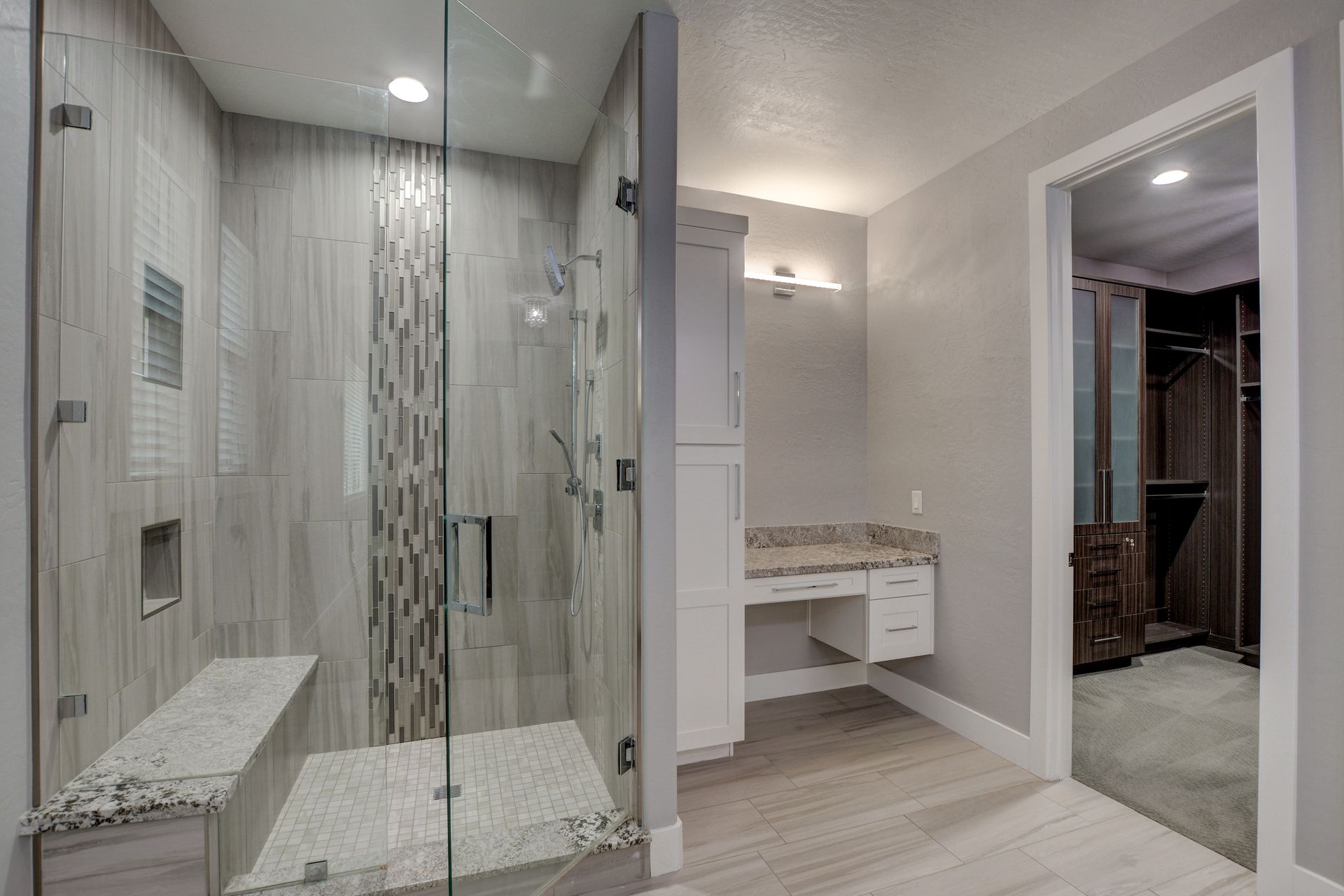Bathroom with glass shower, white cabinets, and vanity. Gray and beige tile with natural light.