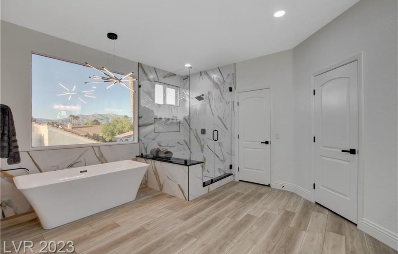 Modern bathroom with large window, freestanding tub, marble shower, and wood-look tile flooring.