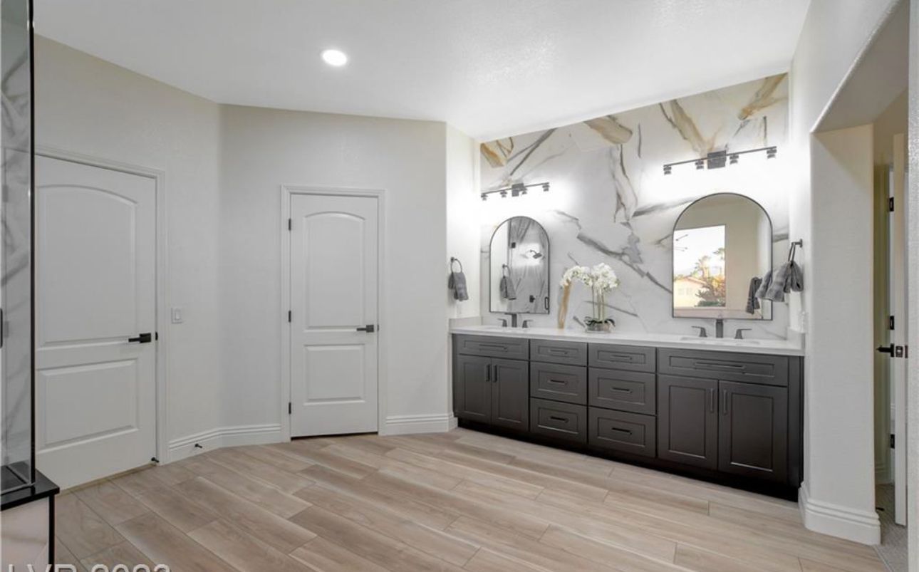 Bathroom with wood-look floor, dark gray cabinets, marble wall, two arched mirrors, and white doors.