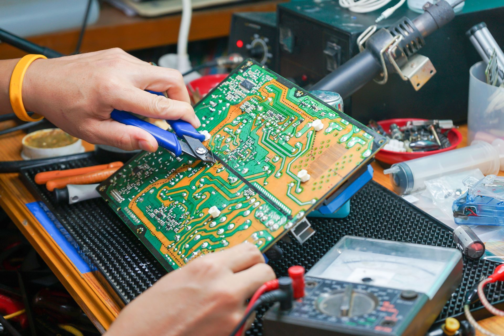 Person using pliers on circuit board at a workbench, with soldering and testing equipment.