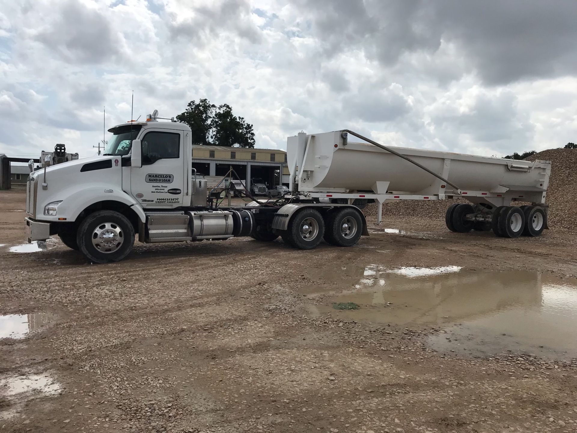 A white semi truck with a dump trailer attached to it is parked in a dirt lot