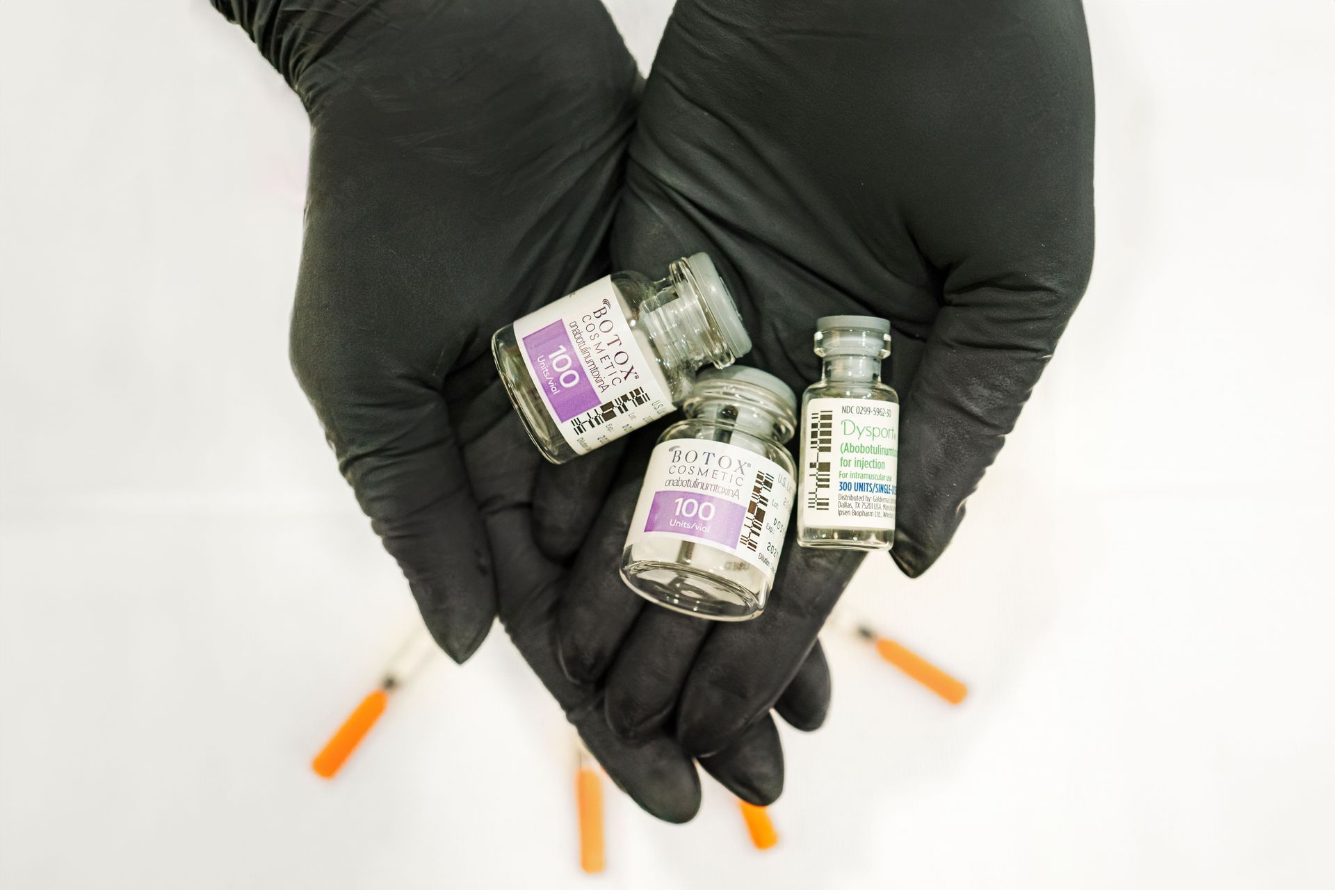 Black-gloved hands holding small medication vials and syringes on a white background