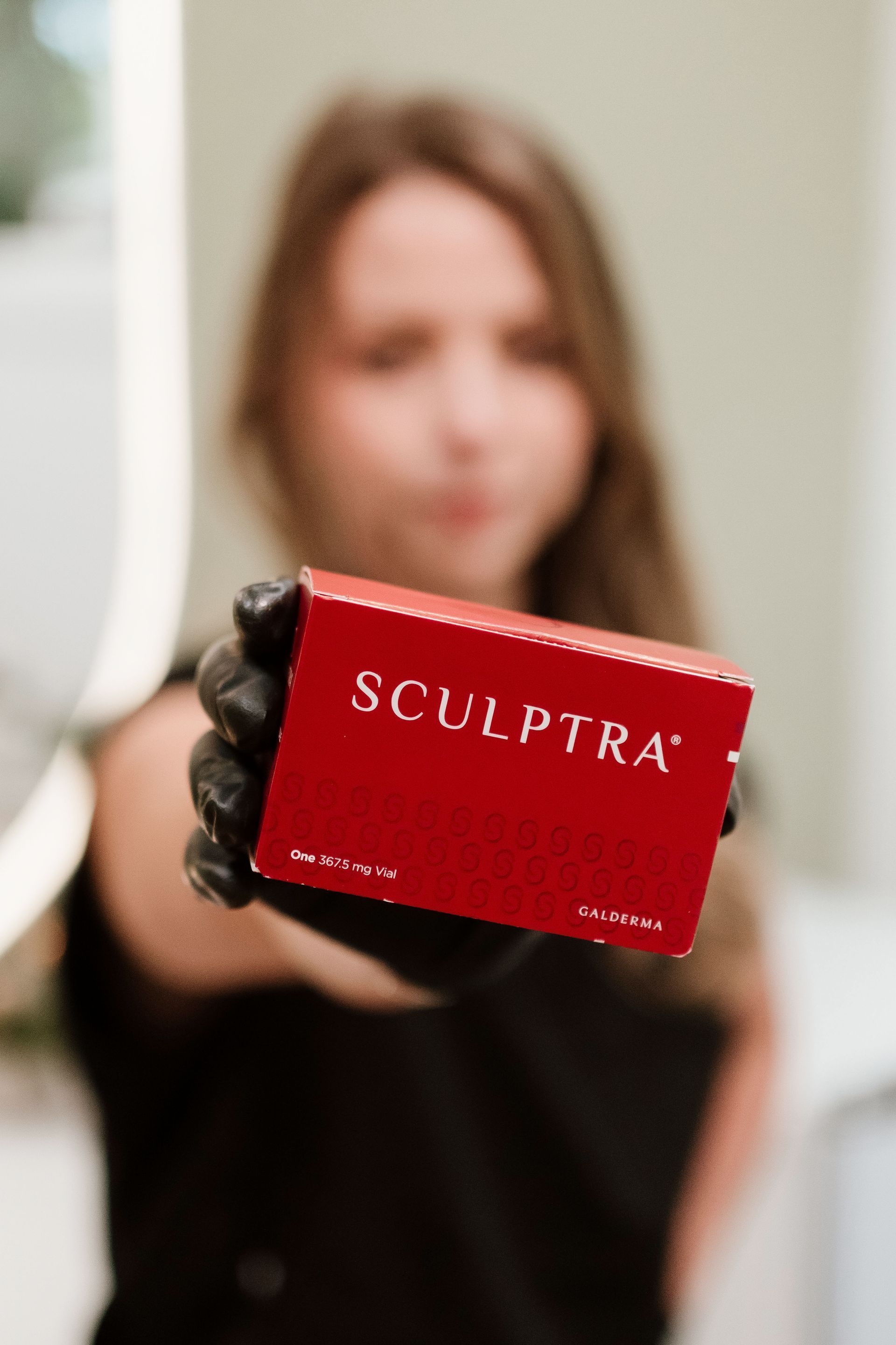 Person holding a red SCULPTRA box toward the camera; blurred background