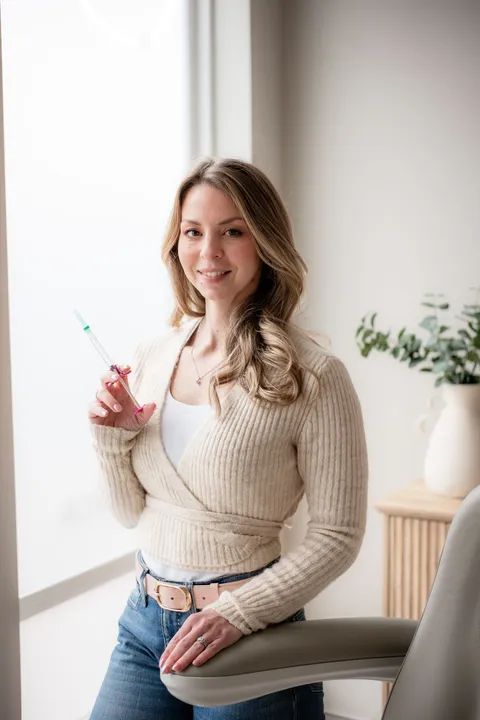 Woman standing by a window, holding a glass with a pink drink, wearing a beige sweater and jeans.