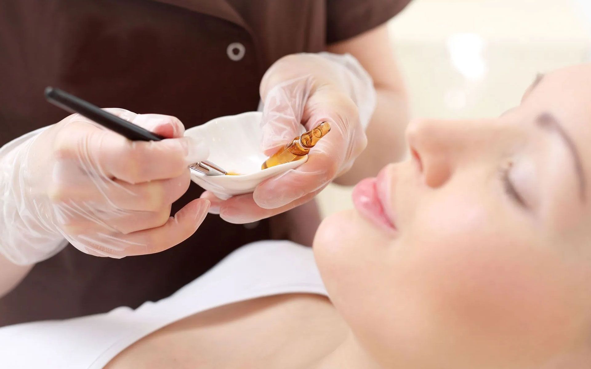 Person applying facial treatment with a small brush while another reclines with eyes closed