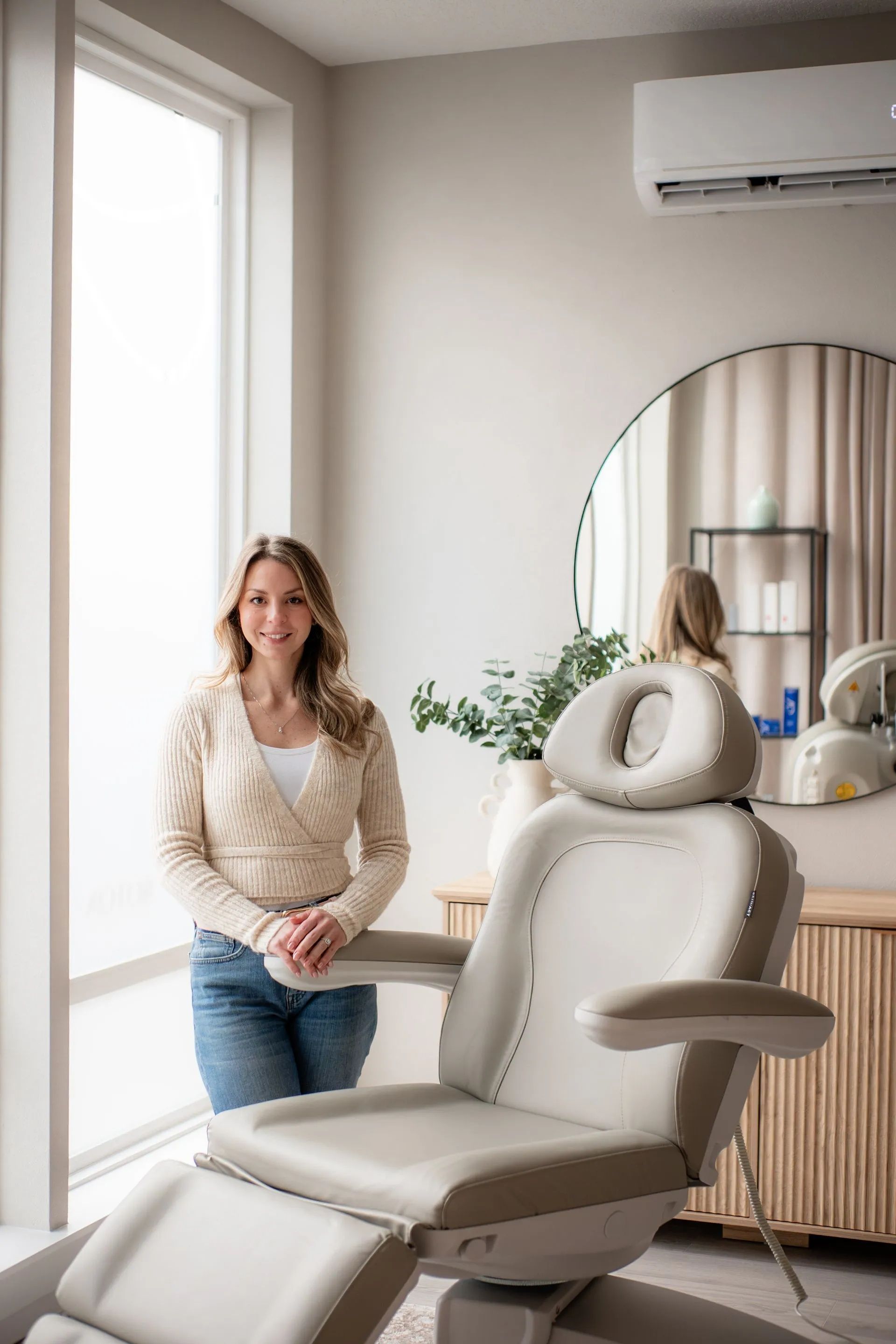 Woman standing beside a gray salon chair in a bright room with a mirror and air conditioner.