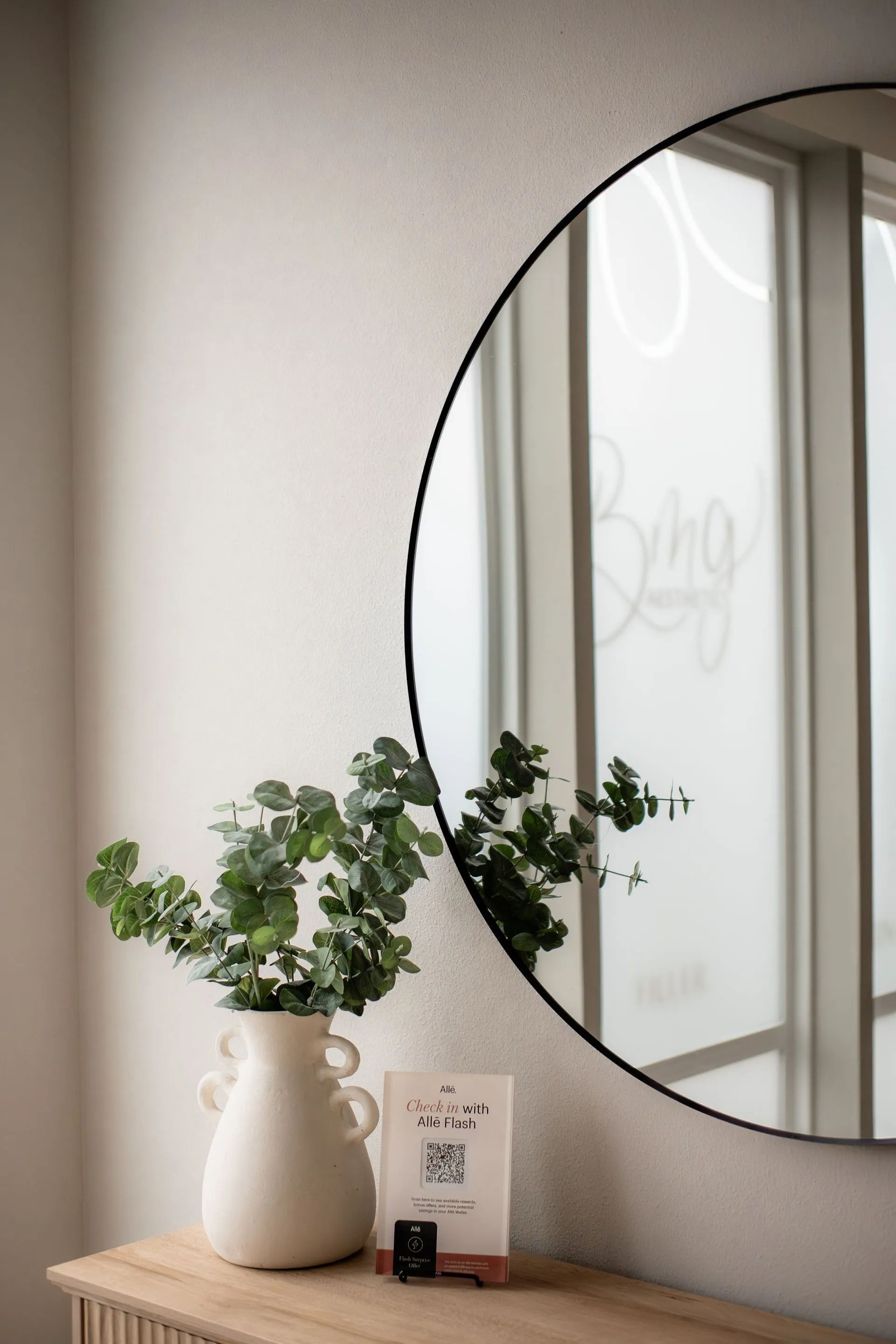 White vase with greenery and a small book on a table beside a round mirror in a softly lit room