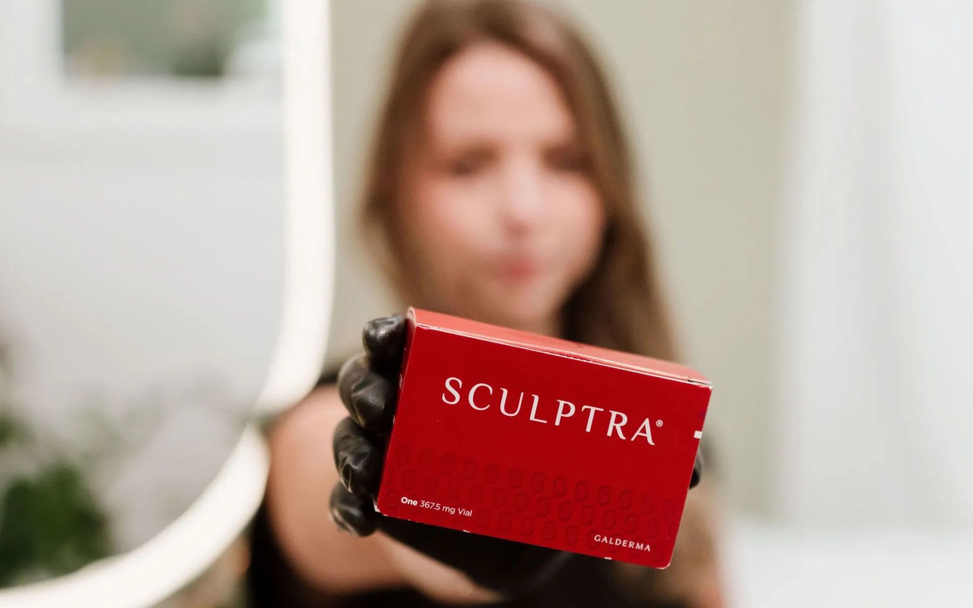 A hand holding a red Sculptra box in front of a blurred person in a bright room