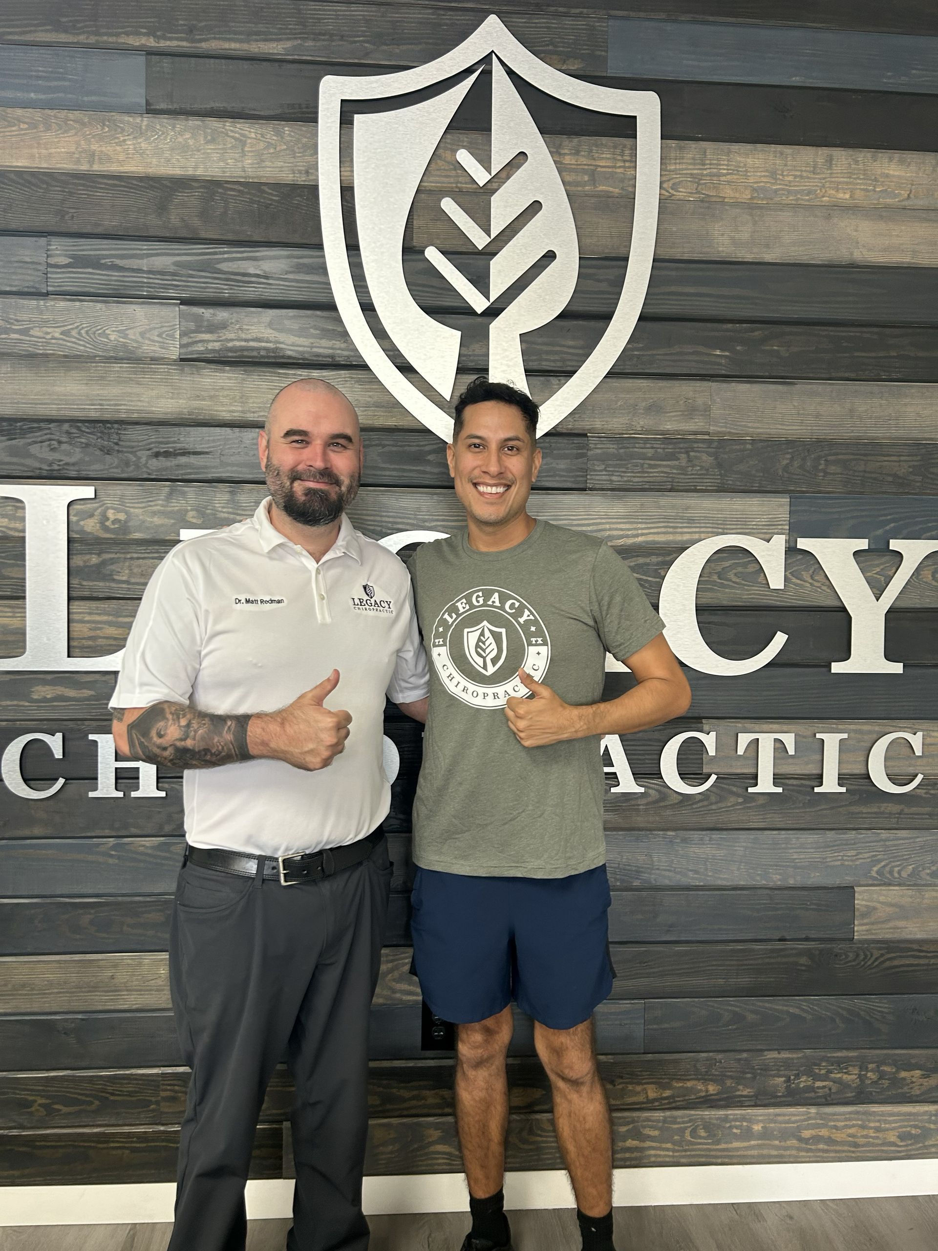 Two men standing side-by-side, smiling and giving a thumbs-up in front of a rustic wood wall with a chiropractic sign.
