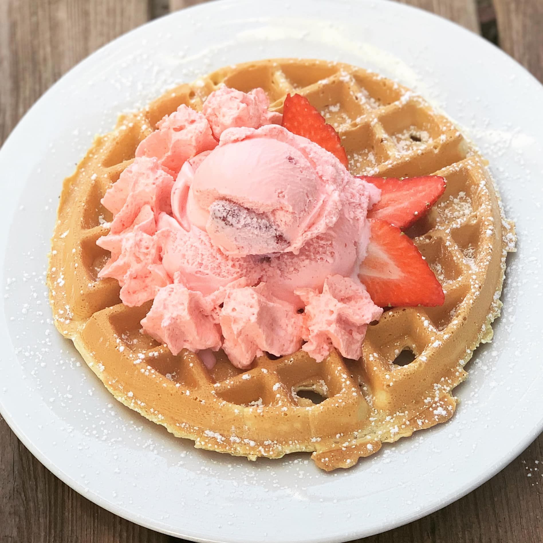 Waffle with pink ice cream, whipped cream, and strawberry slices on a white plate.
