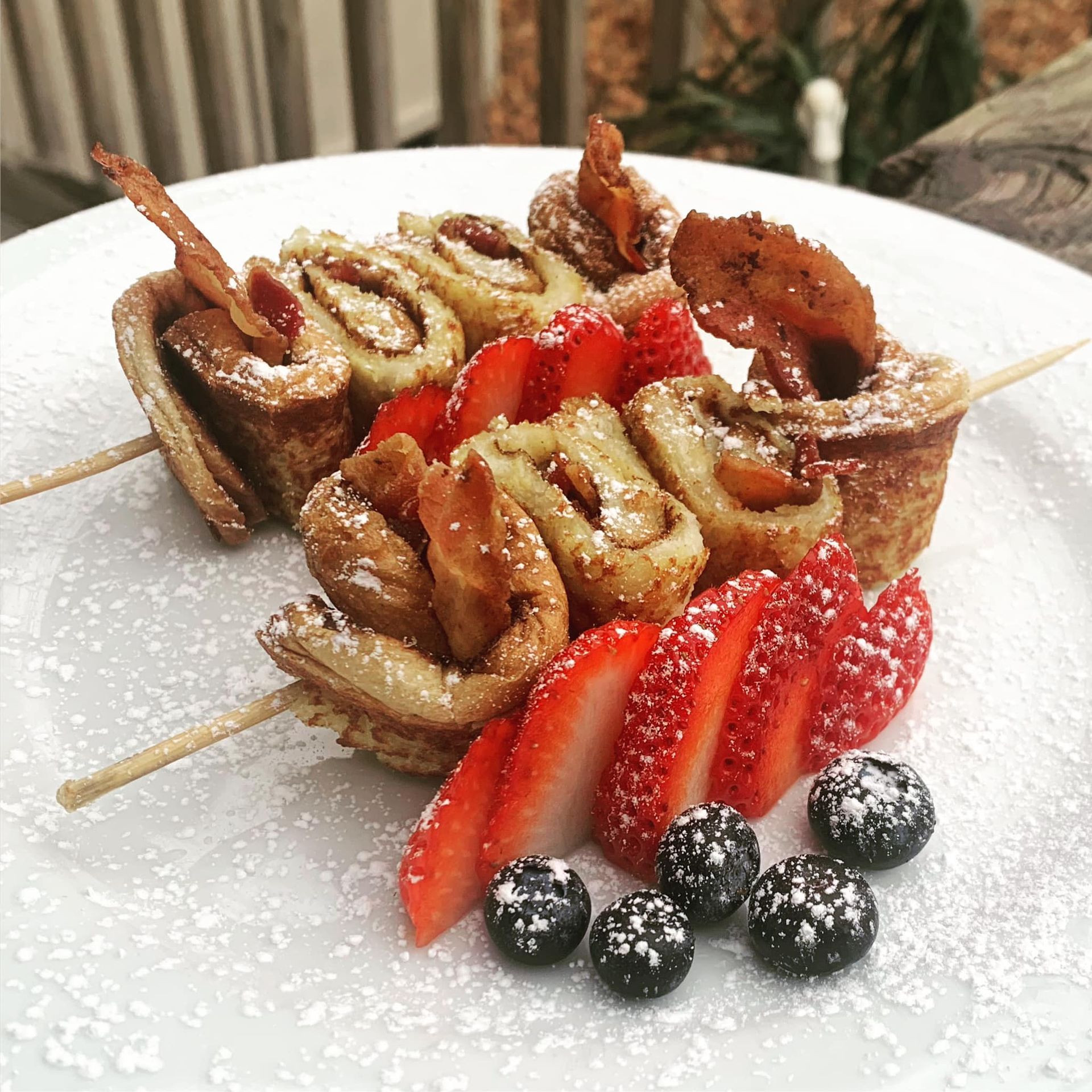 Breakfast skewers with rolled pancakes, bacon, strawberries, blueberries, and powdered sugar.