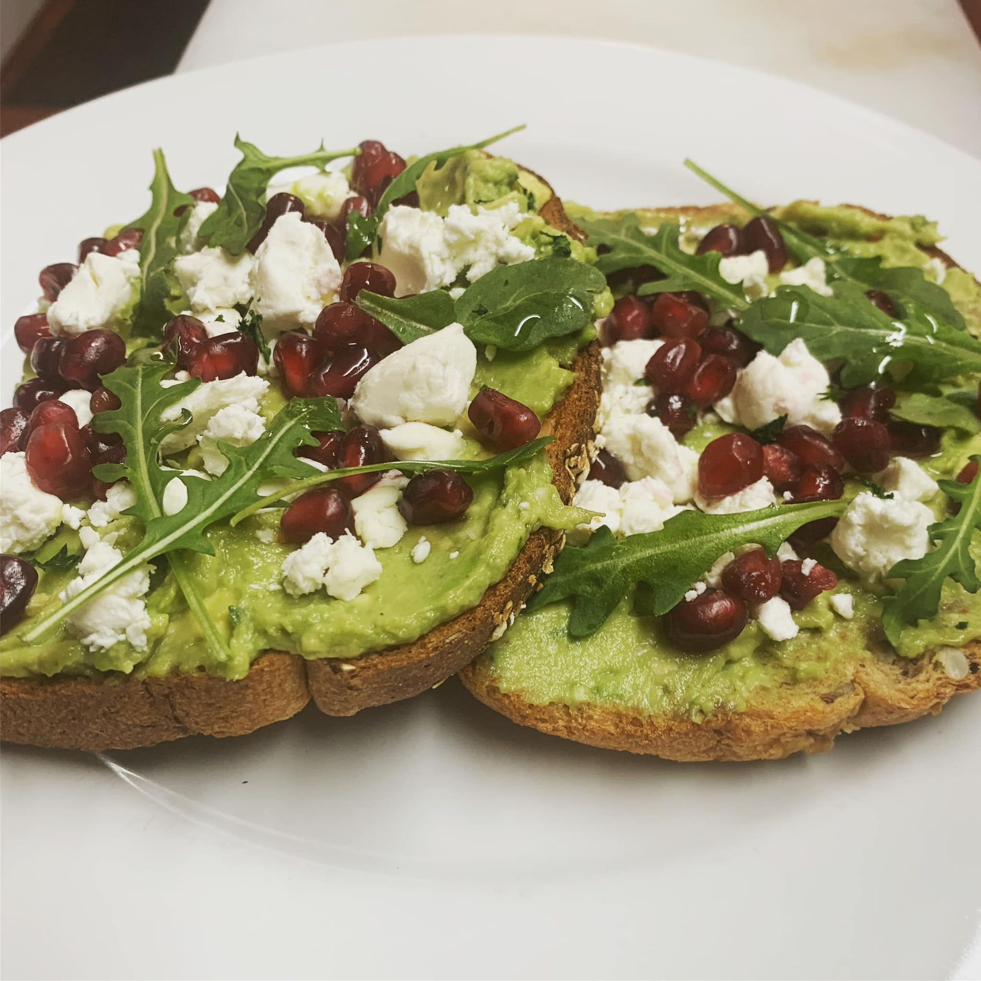 Avocado toast with pomegranate, arugula, and feta on a white plate.