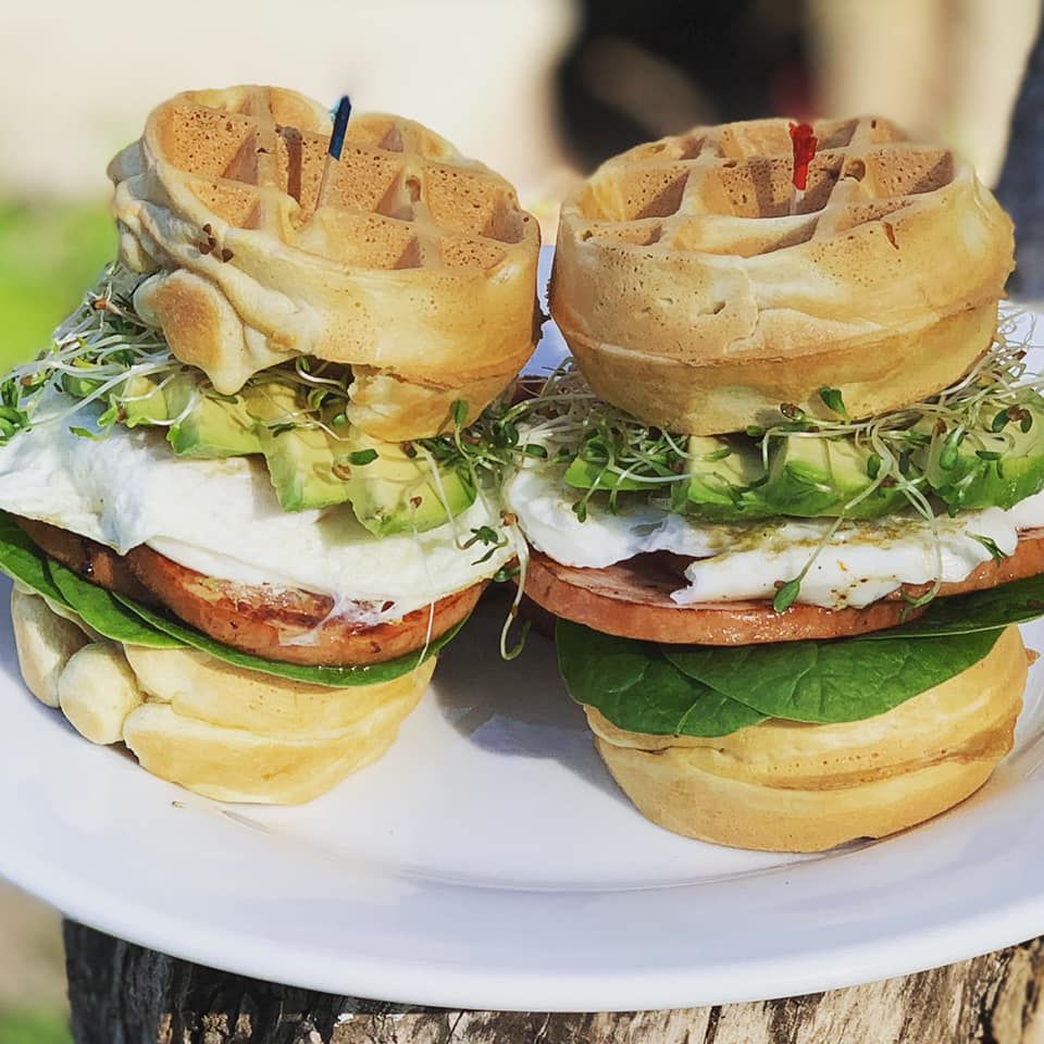 Two breakfast sandwiches on a white plate. Waffle buns, egg, ham, avocado, sprouts, and spinach.