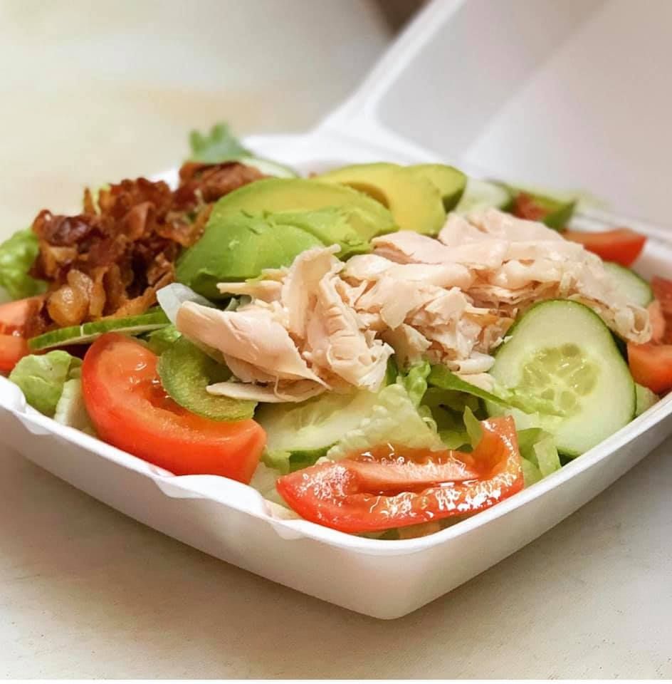 Salad with avocado, tomato, cucumber, bacon, and chicken in a white takeout container.