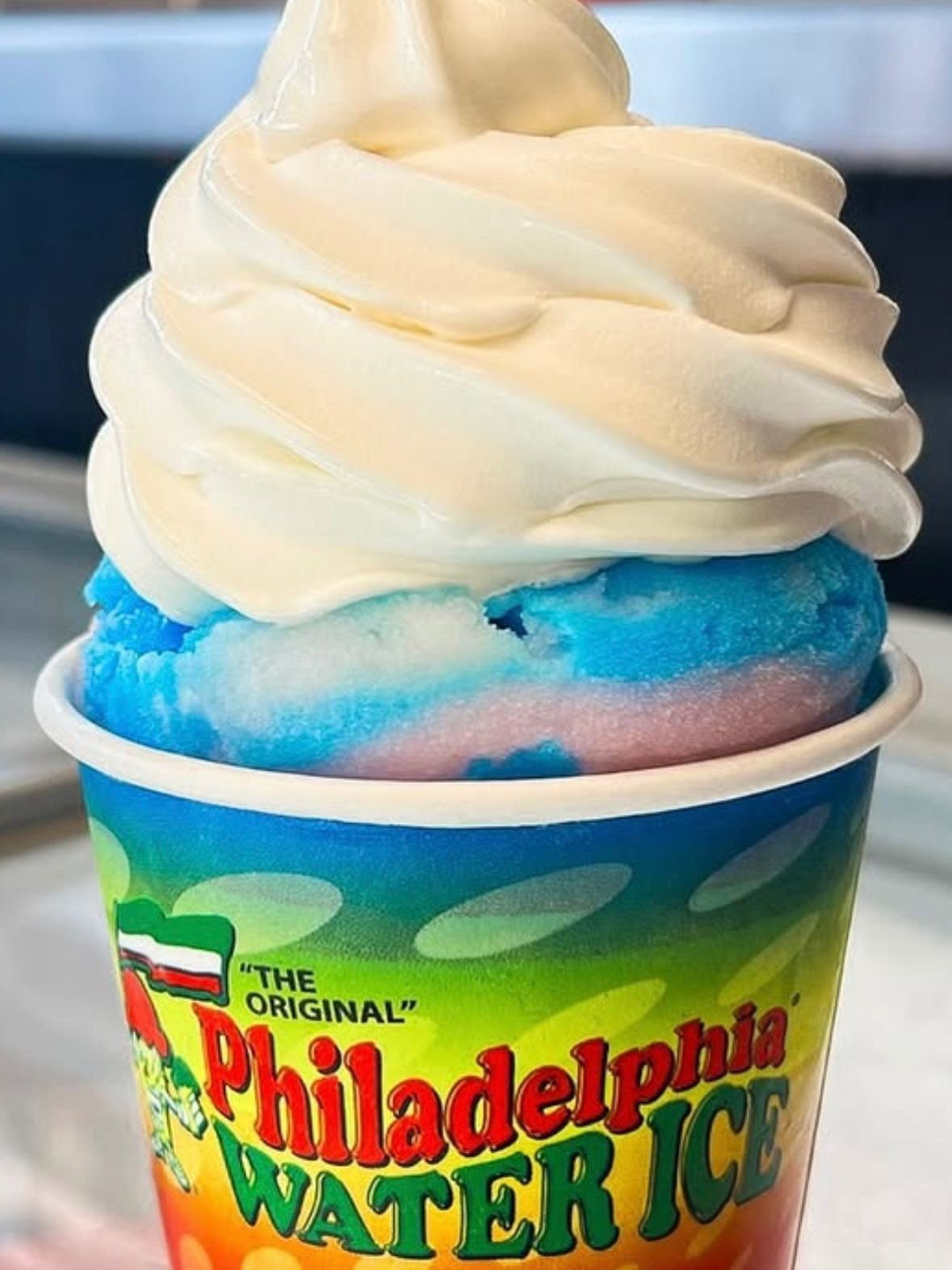 Soft-serve ice cream in a Philadelphia Water Ice cup, with blue and pink layers underneath.