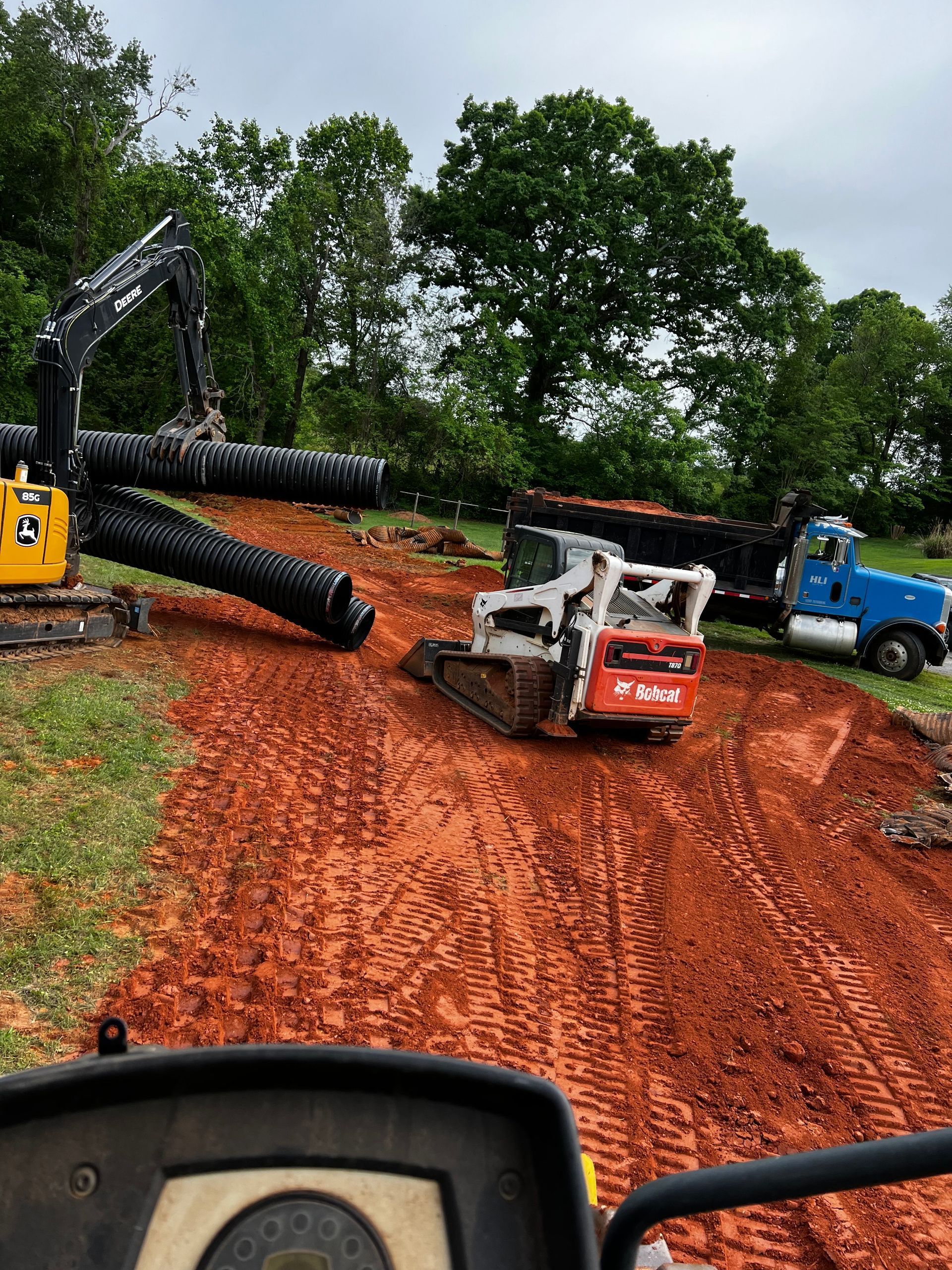 Construction site: Excavator and Bobcat track loader moving pipes on red dirt. Blue truck in the background.