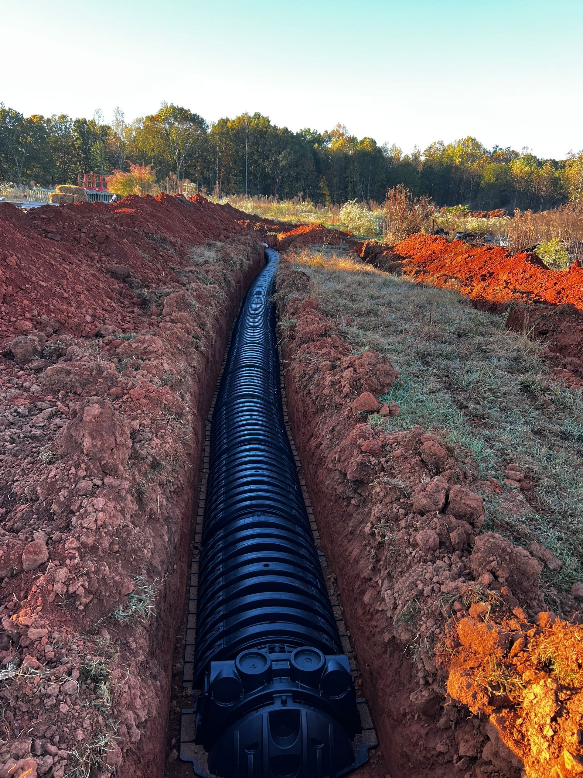 A black corrugated drainage pipe installed in a trench. Red soil on either side, trees in the background.