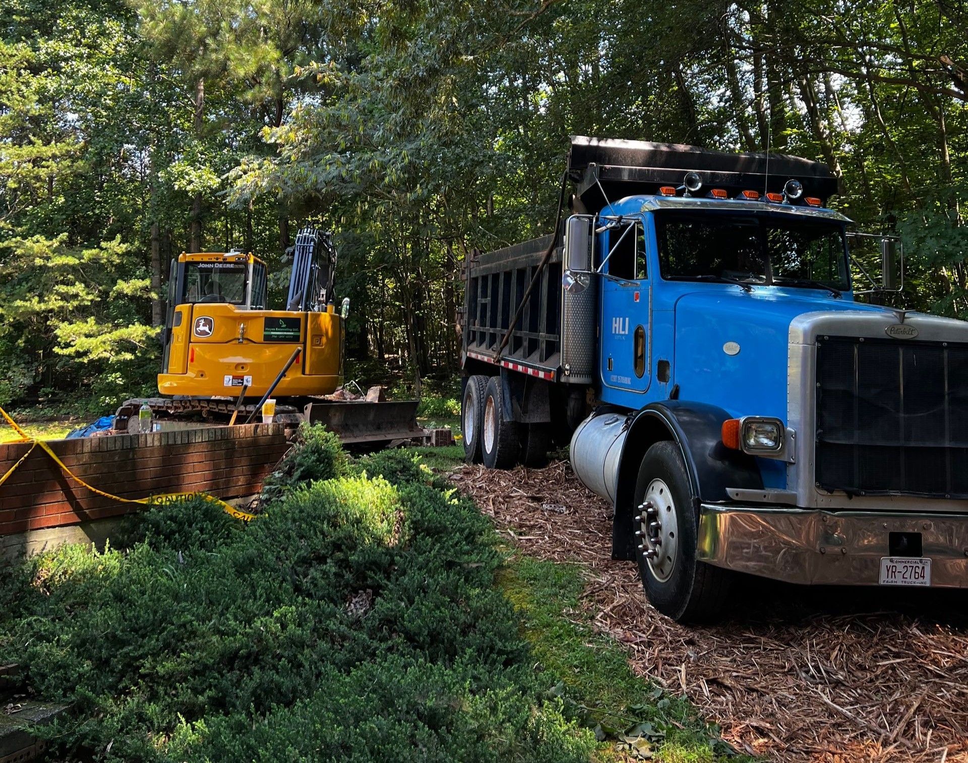 Yellow excavator and blue truck in a wooded area, likely for construction or landscaping work.