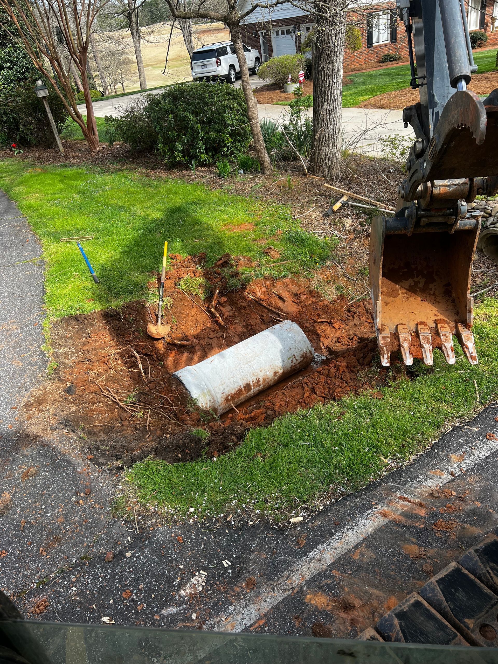 Excavator digging near a sidewalk, exposing a concrete pipe and a yellow utility line. Green grass and trees are in the background.
