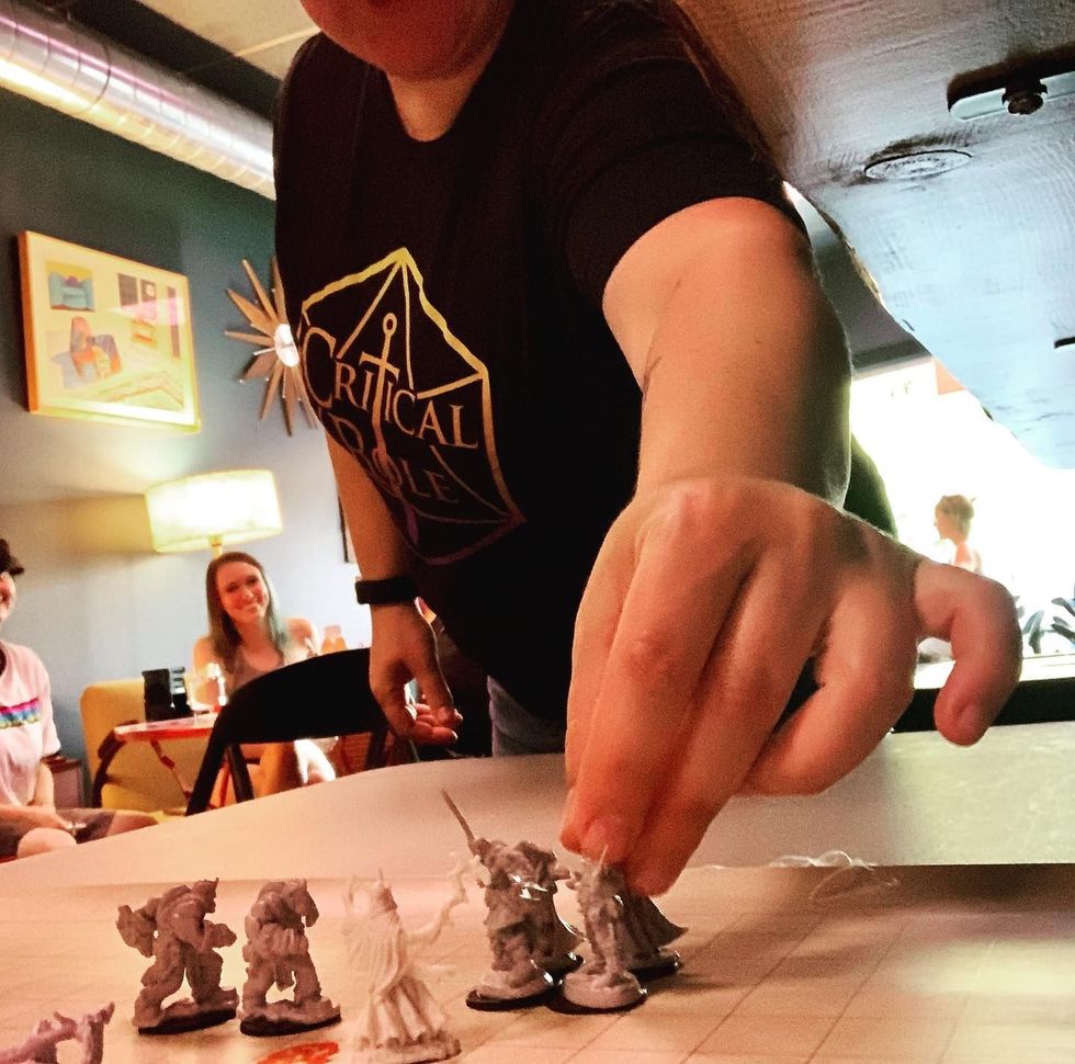 Person sets miniature figures on a tabletop game map. Others watch from a cafe setting.