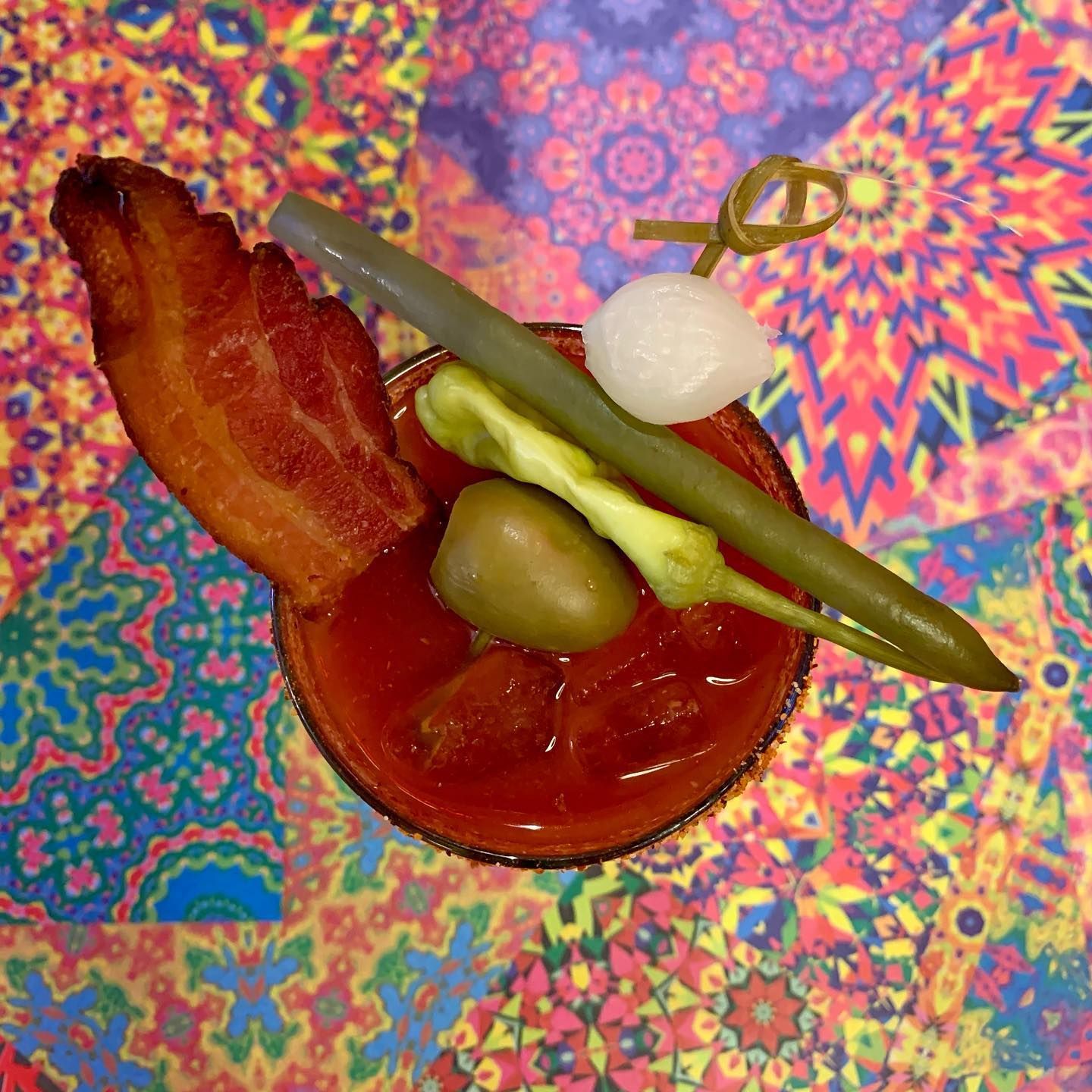 Bloody Mary cocktail with bacon, pickled vegetables, and a toothpick garnish on patterned background.