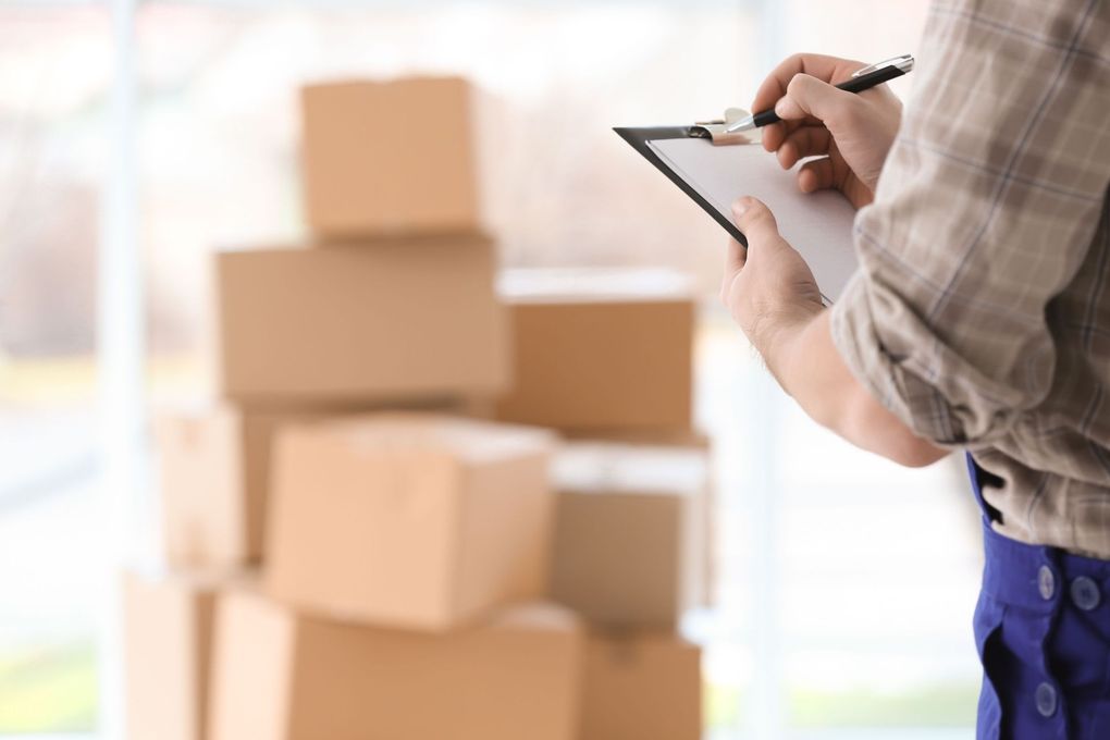 A man is writing on a clipboard in front of a stack of cardboard boxes.