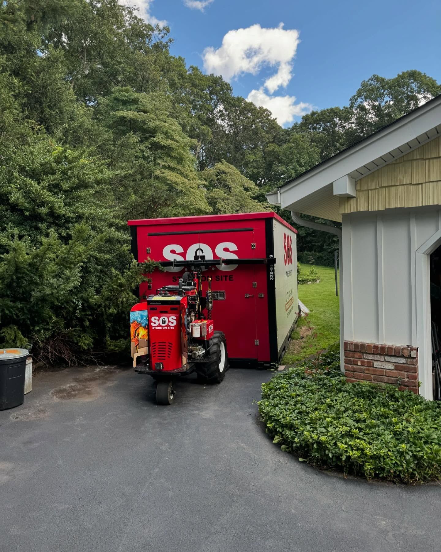Red dumpster pulled by small red machine on a driveway next to a building and trees.