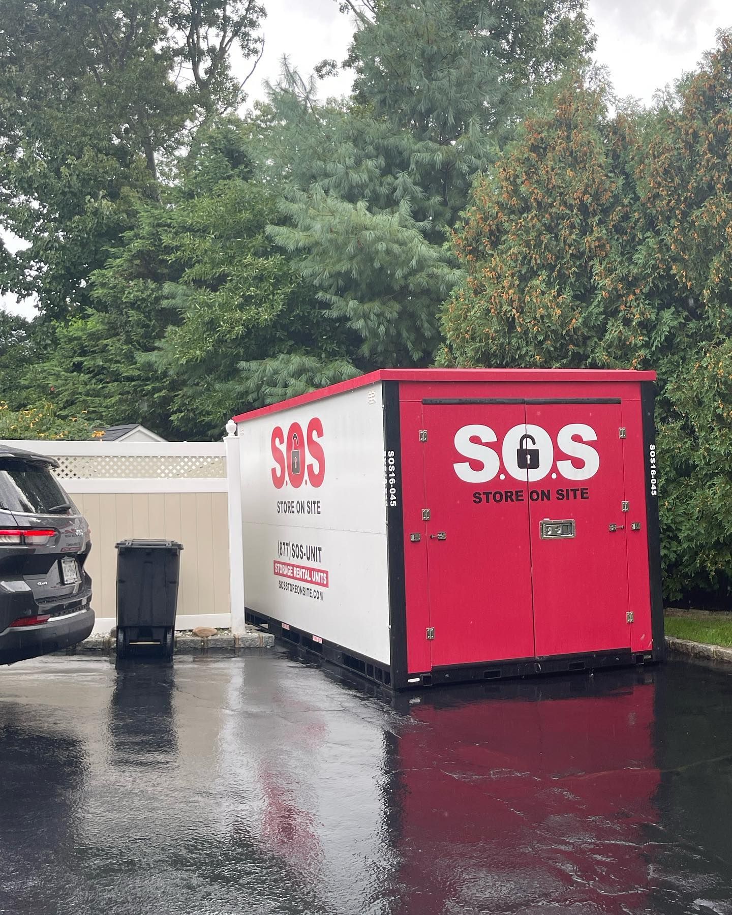 Red and white SOS storage container on wet pavement; parked near a car and bushes.