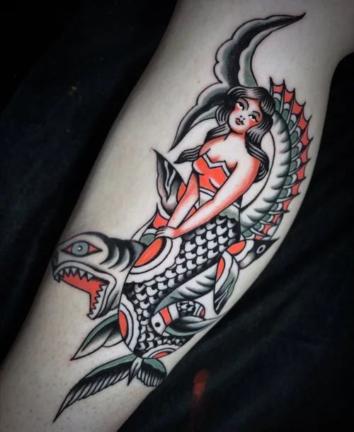 Mermaid tattoo in red, black, and gray riding a fish, with bold tribal-style details