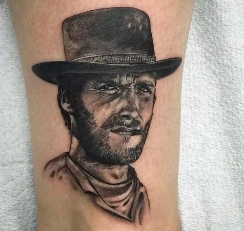 Black-and-gray tattoo of a cowboy with a hat, beard, and neckerchief, winking on skin.