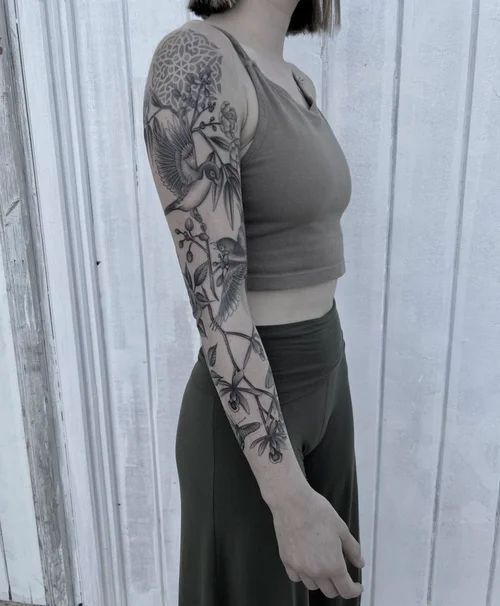Person in a sleeveless top and dark pants showing a floral arm tattoo against a light wall.